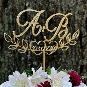 May include: Gold wedding cake topper with the letters A and B intertwined, with a heart between them that says 10.17.2022. The topper is decorated with leafy branches.