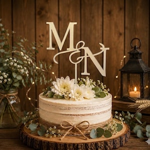 May include: A round cake with light-colored frosting, decorated with white flowers and green leaves, features a twine bow around the base. A gold-colored cake topper displays the letters M and N connected by an ampersand symbol on top.