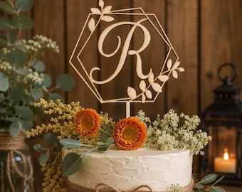 May include: A light-colored wooden cake topper featuring a large, elegant script letter R centered within a geometric hexagonal frame. The frame is decorated with delicate laser-cut leaf motifs on the top left and bottom right corners.