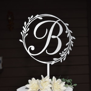 May include: Silver cake topper with a cursive letter B in a circle surrounded by a leafy wreath. The topper is on a white cake with white flowers.