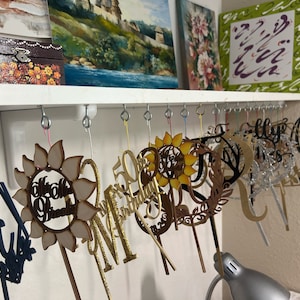 May include: A collection of wooden cake toppers hanging on hooks. The toppers are various shapes and sizes, including a sunflower, a wreath, a letter "M", and a number "50". Some of the toppers are decorated with glitter.