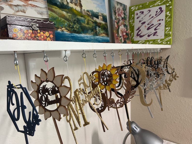 May include: A collection of wooden cake toppers hanging on hooks. The toppers are various shapes and sizes, including a sunflower, a wreath, a letter "M", and a number "50". Some of the toppers are decorated with glitter.