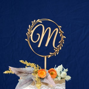 May include: Wooden cake topper with a circle design and the letter "M" in a cursive font. The circle is surrounded by a leafy design. The topper is on a blue background.