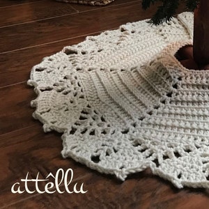 May include: A white crocheted Christmas tree skirt with a lacy design. The skirt is placed on a wooden floor.