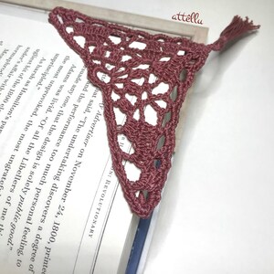 May include: A crocheted bookmark in a dusty rose color with a tassel. The bookmark is shaped like a triangle and has a delicate lace pattern.
