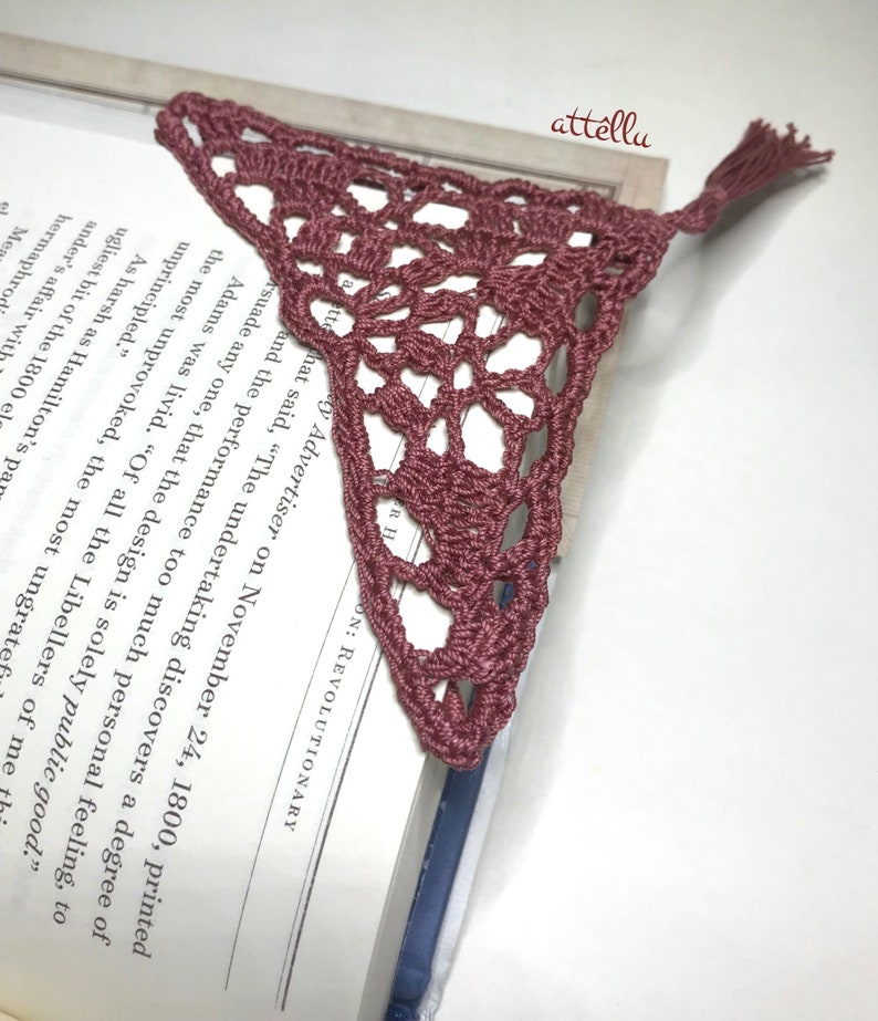 May include: A crocheted bookmark in a dusty rose color with a tassel. The bookmark is shaped like a triangle and has a delicate lace pattern.