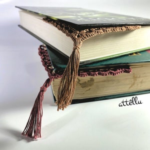 May include: Two books stacked on top of each other with a brown crocheted bookmark with a tassel. The bookmark is on the top book. The bottom book has a burgundy crocheted bookmark with a tassel.