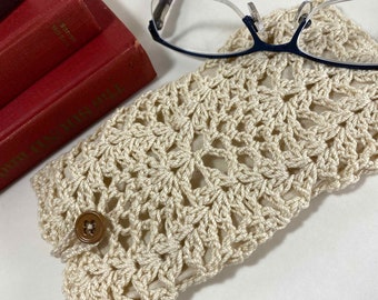 May include: A crocheted beige eyeglass case with a button closure. The case is made with a delicate stitch pattern and has a simple, elegant design.