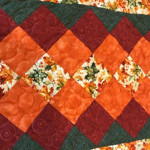May include: A quilted table runner with a diamond pattern in shades of orange, green, and brown. The runner features a fall leaf print with a white background.