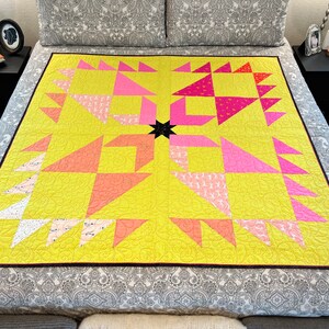 May include: A yellow quilt with a black star in the center. The quilt is made up of pink, orange, and white triangles. The quilt is on a bed with a gray and white patterned bedspread.