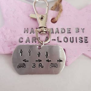 May include: A silver keychain with a small metal tag that has the words "Handmade by Carla-Louise" and a gamepad with directional arrows and the letters "BA" engraved on it.