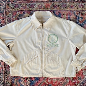 Może przedstawiać: Kremowa kurtka zapinana na zamek błyskawiczny z dwoma kieszeniami. Kurtka ma haftowany zielono-biały logo z napisem "WAYNE".