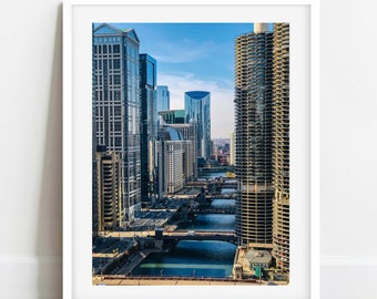 May include: A high-angle view of a cityscape with tall buildings and a river running through the middle. The buildings are mostly glass and steel, and the river is a deep blue colour. The sky is a bright blue, and there are some clouds in the distance.