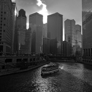 K&ouml;nnte beinhalten: Ein Schwarzwei&szlig;foto einer Skyline mit hohen Geb&auml;uden und einem Fluss im Vordergrund. Ein Boot mit Menschen f&auml;hrt auf dem Fluss.