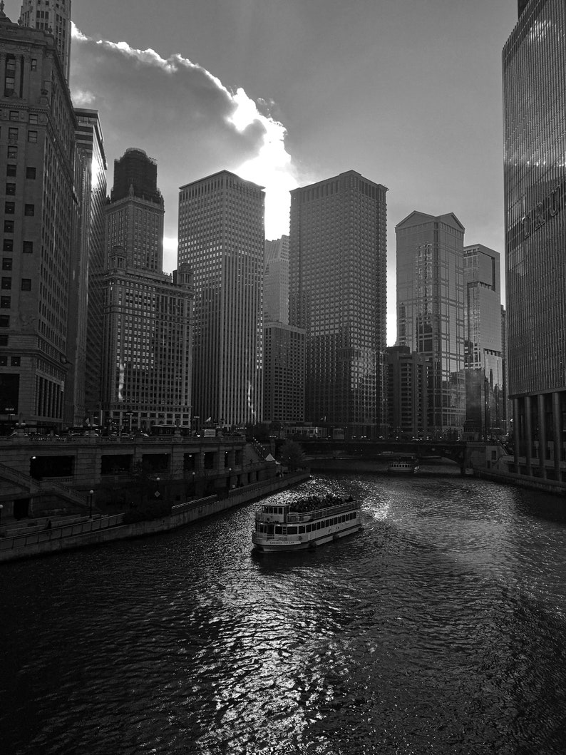 K&ouml;nnte beinhalten: Ein Schwarzwei&szlig;foto einer Skyline mit hohen Geb&auml;uden und einem Fluss im Vordergrund. Ein Boot mit Menschen f&auml;hrt auf dem Fluss.