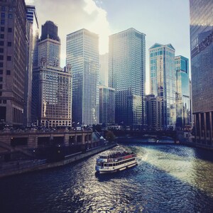 K&ouml;nnte beinhalten: Ein Boot mit Passagieren f&auml;hrt auf einem Fluss in Chicago. Der Fluss ist von hohen Geb&auml;uden ges&auml;umt. Der Himmel ist blau mit wei&szlig;en Wolken.