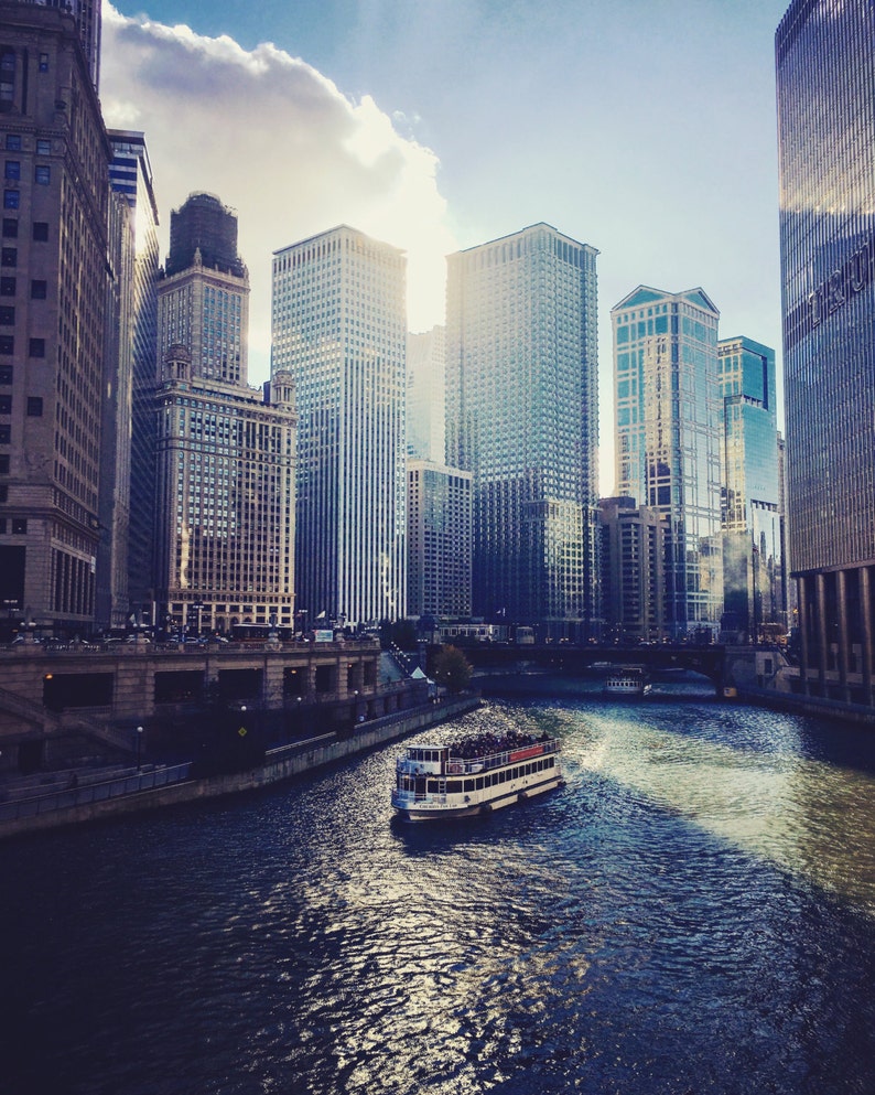 K&ouml;nnte beinhalten: Ein Boot mit Passagieren f&auml;hrt auf einem Fluss in Chicago. Der Fluss ist von hohen Geb&auml;uden ges&auml;umt. Der Himmel ist blau mit wei&szlig;en Wolken.