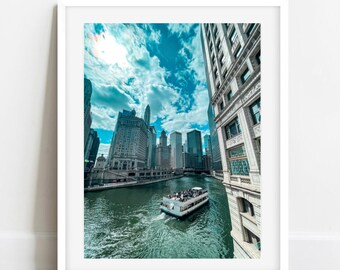 May include: A white boat with people on board sails down a river in Chicago, Illinois. The boat is surrounded by tall buildings and a blue sky with white clouds.