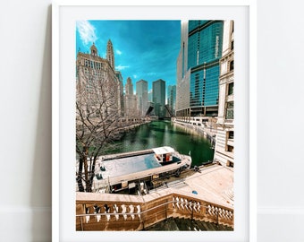 May include: A framed print depicting a cityscape with a river, buildings, and a boat. The image has a bright blue sky and a bridge in the background. The print is displayed on a wooden floor.