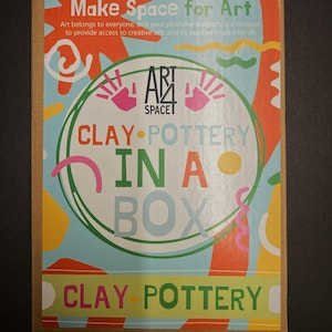 Puede incluir: Una caja de cartón con un diseño colorido que promociona "Clay Pottery in a Box". La caja presenta el texto "Make Space for Art" y "Art Space". El diseño incluye huellas de manos y formas abstractas en naranja, rosa y azul.