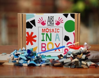 Puede incluir: Una caja de cartón con el texto "Art Space 1 Mosaic in a Box" impreso en ella. La caja contiene una variedad de azulejos de mosaico de colores, un pincel y un pequeño recipiente de pegamento.