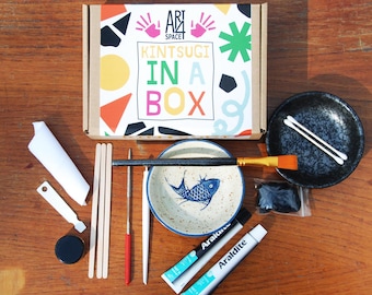 Könnte beinhalten: Kintsugi-Set in einer Box mit einer kleinen blauen und weißen Keramikschale mit Fischdesign, zwei Klebstoffröhrchen, einer kleinen schwarzen Schale, zwei Wattestäbchen, einem kleinen schwarzen Deckel, einem kleinen schwarzen Behälter, einem kleinen weißen Behälter, einem kleinen roten Werkzeug, einem kleinen weißen Werkzeug, einem kleinen schwarzen Werkzeug, einem kleinen weißen Werkzeug, drei Holzstäbchen und einem kleinen Pinsel.