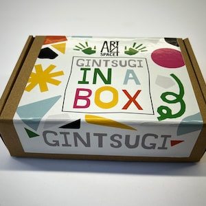 May include: A brown cardboard box with a colorful lid featuring the text "GINTUSGI IN A BOX". The lid has a white background with various shapes and the words "ABI SPACE". The box is closed.