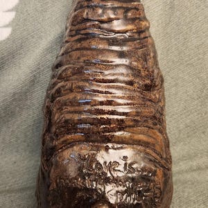 May include: A brown ceramic sculpture with a glossy finish and horizontal ridged texture. The surface features the embossed text "Love is..." above a small triangular cluster of circular shapes, creating a tactile and layered appearance.
