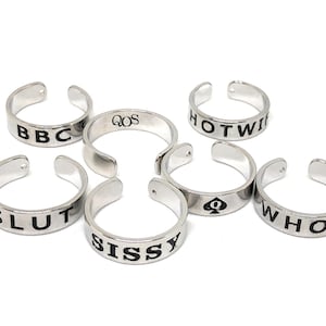 May include: Six silver rings with black lettering. The rings say "BBC", "SLUT", "SISSY", "QOS", "HOTWIFE", and "WHORE".