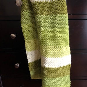 Może przedstawiać: Ręcznie robiony szalik z wzorem ściegu o fakturze. Szalik ma poziome paski w kolorze białym i r&oacute;żnych odcieniach zieleni. Szalik jest narzucony na ciemnobrązową komodę.