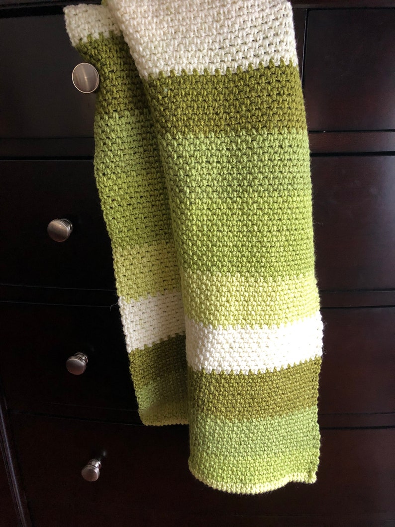 Może przedstawiać: Ręcznie robiony szalik z wzorem ściegu o fakturze. Szalik ma poziome paski w kolorze białym i r&oacute;żnych odcieniach zieleni. Szalik jest narzucony na ciemnobrązową komodę.