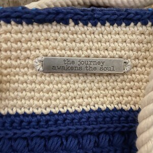Puede incluir: Primer plano de un bolso tote tejido a ganchillo con un patr&oacute;n de rayas crema y azul marino. El bolso tiene un asa de cuerda y una peque&ntilde;a etiqueta met&aacute;lica que dice "the journey awakens the soul."