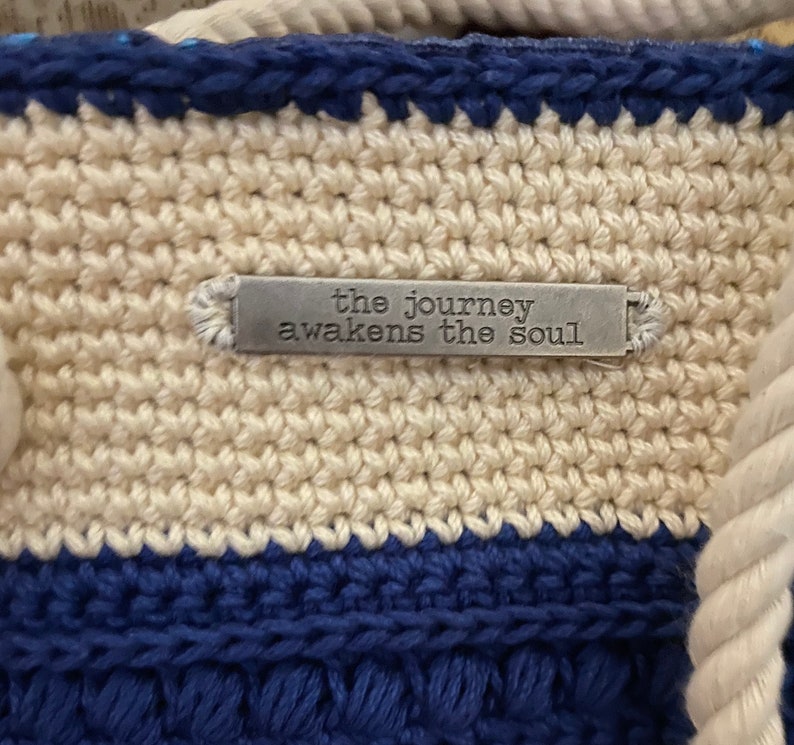 Puede incluir: Primer plano de un bolso tote tejido a ganchillo con un patr&oacute;n de rayas crema y azul marino. El bolso tiene un asa de cuerda y una peque&ntilde;a etiqueta met&aacute;lica que dice "the journey awakens the soul."