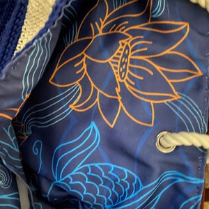 Puede incluir: Bolsa azul con cord&oacute;n y dise&ntilde;o floral. El interior presenta una gran flor de loto naranja y patrones de escamas de pez azules sobre un fondo azul oscuro. El exterior de la bolsa es de tejido azul tejido con ribete blanco.