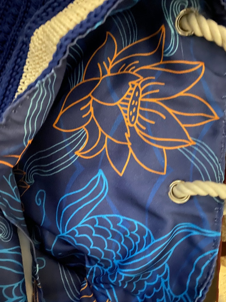 Puede incluir: Bolsa azul con cord&oacute;n y dise&ntilde;o floral. El interior presenta una gran flor de loto naranja y patrones de escamas de pez azules sobre un fondo azul oscuro. El exterior de la bolsa es de tejido azul tejido con ribete blanco.
