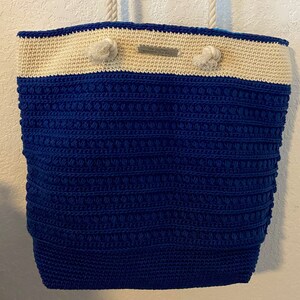 Puede incluir: Un bolso tote grande, hecho a mano, con un cuerpo de ganchillo azul marino texturizado y una secci&oacute;n superior de color crema. El bolso presenta asas de cuerda y nudos decorativos. Una peque&ntilde;a etiqueta met&aacute;lica est&aacute; adherida en la parte superior. El bolso est&aacute; dise&ntilde;ado para transportar objetos.