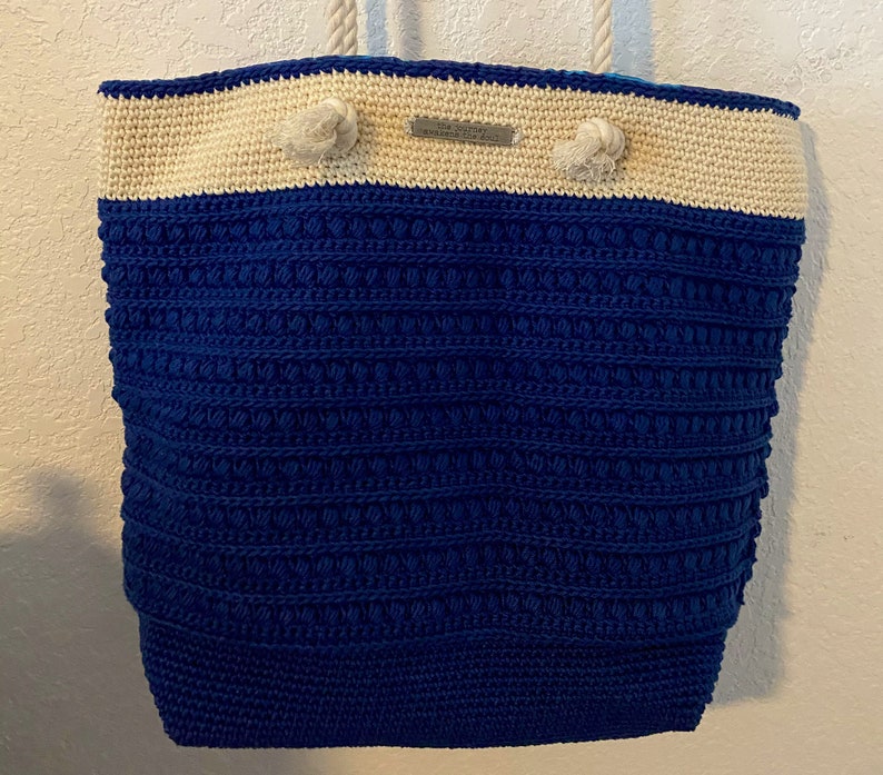 Puede incluir: Un bolso tote grande, hecho a mano, con un cuerpo de ganchillo azul marino texturizado y una secci&oacute;n superior de color crema. El bolso presenta asas de cuerda y nudos decorativos. Una peque&ntilde;a etiqueta met&aacute;lica est&aacute; adherida en la parte superior. El bolso est&aacute; dise&ntilde;ado para transportar objetos.