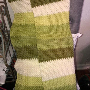 Może przedstawiać: Ręcznie robiona, pasiasta chusta w odcieniach zieleni i kremu. Chusta wykonana jest z teksturowanego, dzianinowego materiału. Paleta kolor&oacute;w obejmuje jasną zieleń, oliwkową zieleń i krem, tworząc przytulną i zachęcającą estetykę. Chusta jest narzucona na krzesło.