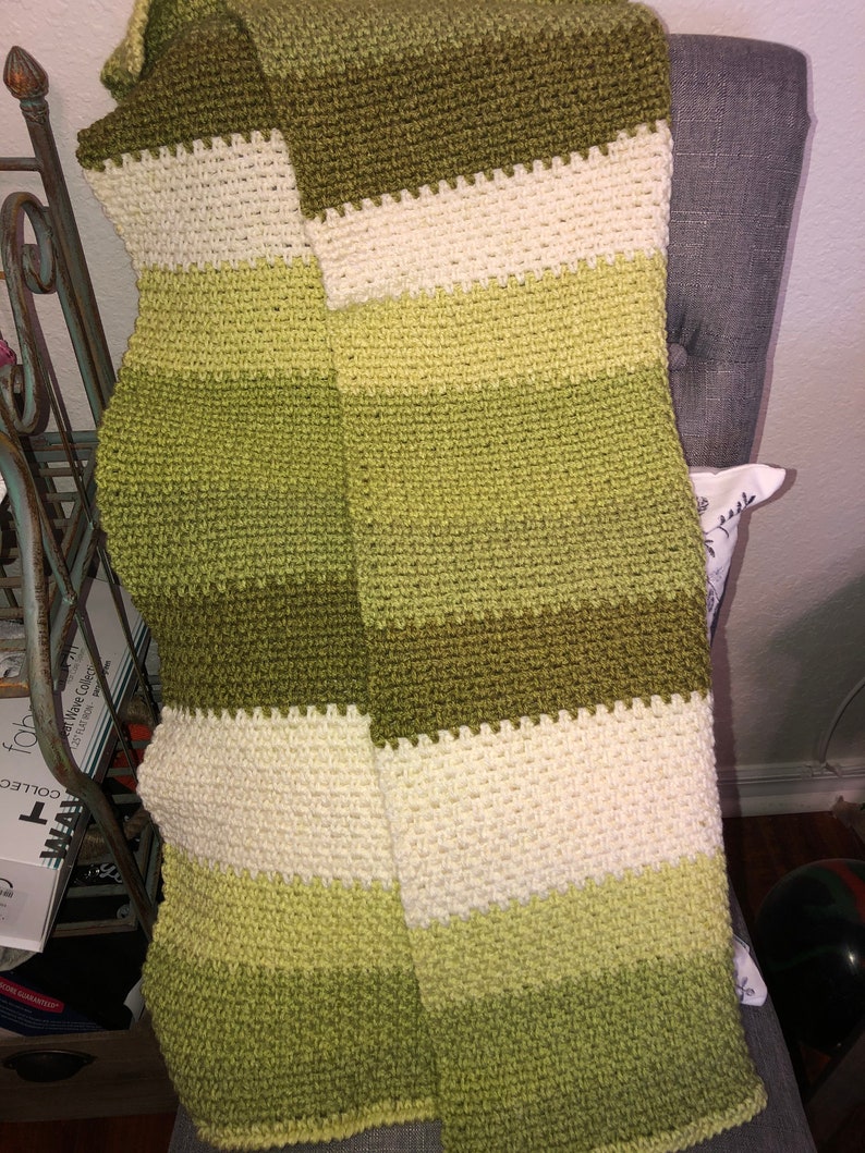 Może przedstawiać: Ręcznie robiona, pasiasta chusta w odcieniach zieleni i kremu. Chusta wykonana jest z teksturowanego, dzianinowego materiału. Paleta kolor&oacute;w obejmuje jasną zieleń, oliwkową zieleń i krem, tworząc przytulną i zachęcającą estetykę. Chusta jest narzucona na krzesło.