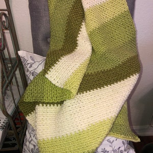 Może przedstawiać: Ręcznie robiony koc z szydełka w odcieniach zieleni i kremu, narzucony na szary fotel. Koc ma poziome paski w r&oacute;żnych odcieniach zieleni i kremu. Widoczna jest faktura ścieg&oacute;w szydełkowych, tworząc przytulny i zachęcający wygląd.