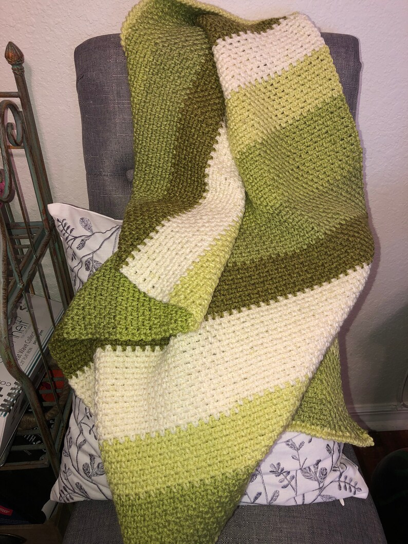 Może przedstawiać: Ręcznie robiony koc z szydełka w odcieniach zieleni i kremu, narzucony na szary fotel. Koc ma poziome paski w r&oacute;żnych odcieniach zieleni i kremu. Widoczna jest faktura ścieg&oacute;w szydełkowych, tworząc przytulny i zachęcający wygląd.