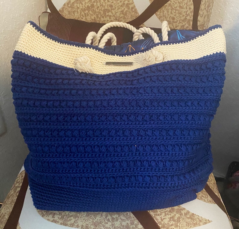 Puede incluir: Un bolso tote grande de ganchillo azul con un borde superior color crema y asas de cuerda. El bolso presenta un patr&oacute;n de puntadas texturizadas y un forro interior estampado. Una peque&ntilde;a etiqueta met&aacute;lica est&aacute; adherida al frente.