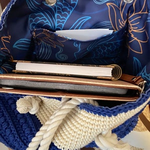 Puede incluir: Un bolso tote de crochet azul y crema con asa de cuerda. El interior presenta una tela azul marino con un estampado floral y contiene un cuaderno y una tableta. El bolso est&aacute; abierto, revelando su contenido.