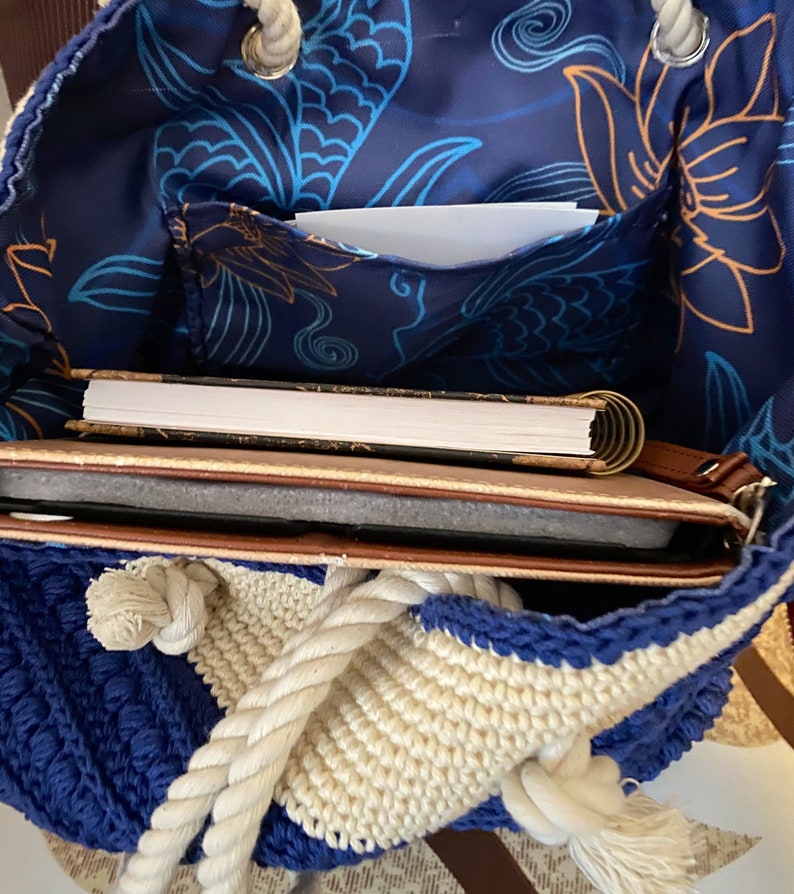 Puede incluir: Un bolso tote de crochet azul y crema con asa de cuerda. El interior presenta una tela azul marino con un estampado floral y contiene un cuaderno y una tableta. El bolso est&aacute; abierto, revelando su contenido.