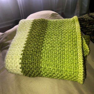 Może przedstawiać: Złożony, ręcznie robiony koc szydełkowy w odcieniach zieleni. Koc ma wzorzysty ścieg i gradient kolor&oacute;w, przechodzący od jasnej zieleni do ciemniejszej zieleni oliwkowej. Krawędzie są starannie wykończone, ukazując kunszt wykonania.