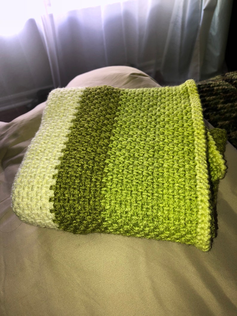 Może przedstawiać: Złożony, ręcznie robiony koc szydełkowy w odcieniach zieleni. Koc ma wzorzysty ścieg i gradient kolor&oacute;w, przechodzący od jasnej zieleni do ciemniejszej zieleni oliwkowej. Krawędzie są starannie wykończone, ukazując kunszt wykonania.