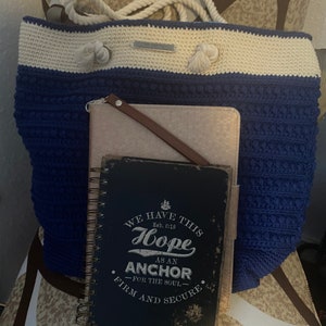 Puede incluir: Un bolso tote de ganchillo azul y crema con asas de cuerda y una peque&ntilde;a etiqueta. Una cartera de cuero rosa claro y un diario con espiral con el texto "WE HAVE THIS HOPE AS AN ANCHOR" est&aacute;n delante del bolso.