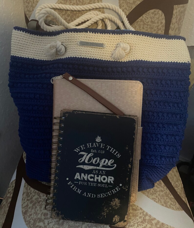 Puede incluir: Un bolso tote de ganchillo azul y crema con asas de cuerda y una peque&ntilde;a etiqueta. Una cartera de cuero rosa claro y un diario con espiral con el texto "WE HAVE THIS HOPE AS AN ANCHOR" est&aacute;n delante del bolso.
