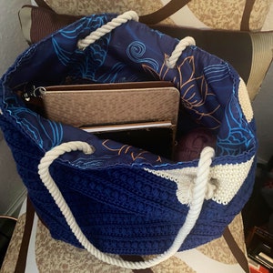 Puede incluir: Un bolso tote de crochet azul hecho a mano con asas de cuerda blanca. El interior de la bolsa presenta una tela azul con un estampado floral y de olas. Dentro de la bolsa hay varios art&iacute;culos, incluyendo un cuaderno marr&oacute;n y una madeja de lana morada.