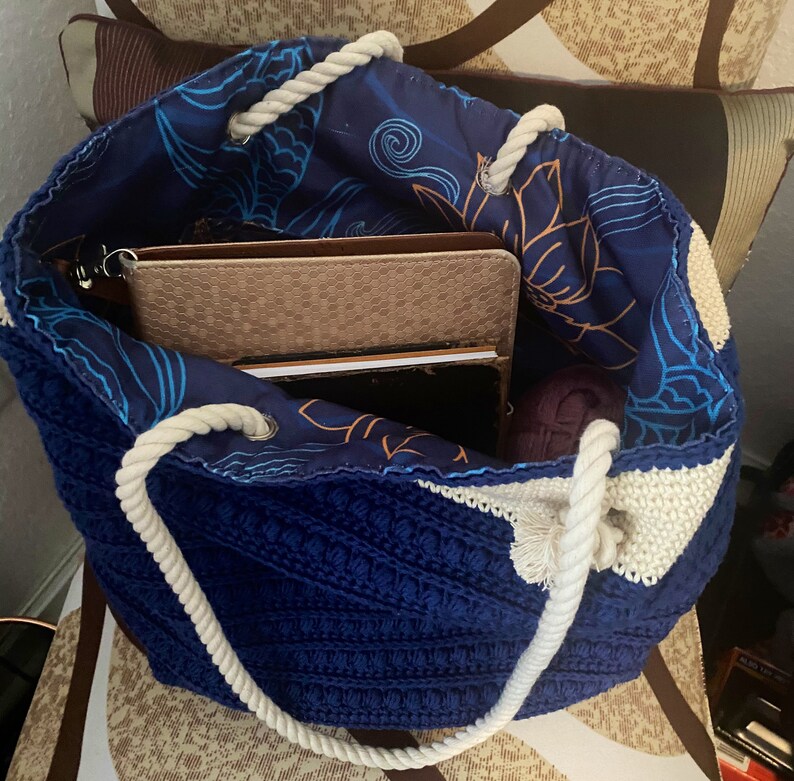 Puede incluir: Un bolso tote de crochet azul hecho a mano con asas de cuerda blanca. El interior de la bolsa presenta una tela azul con un estampado floral y de olas. Dentro de la bolsa hay varios art&iacute;culos, incluyendo un cuaderno marr&oacute;n y una madeja de lana morada.